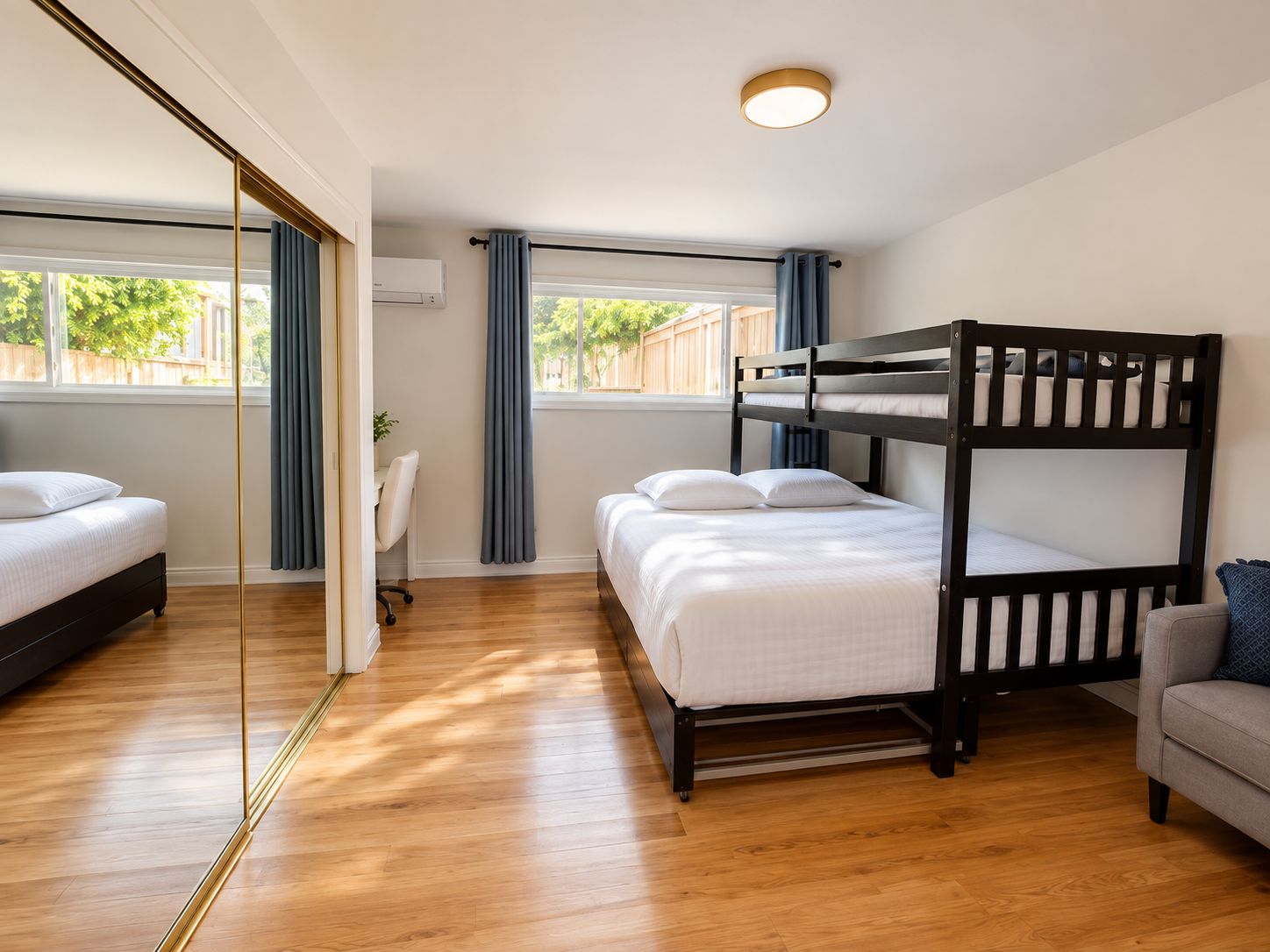 A room with a bunk bed, a desk, and a chair. There is a window with curtains and a view of trees outside.