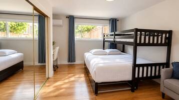 A room with a bunk bed, a desk, and a chair. There is a window with curtains and a view of trees outside.