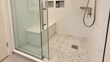 Un baño moderno con ducha envuelta en vidrio, paredes de azulejos blancos y un banco integrado.