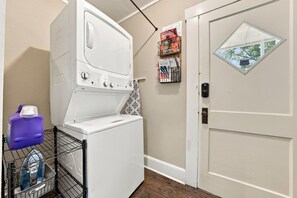 A laundry room with a front-loading washer and dryer, a basket of laundry, and a door with a window. - Violet Haus - 3 blocks from downtown (Fredericksburg)
