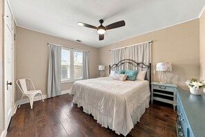A bedroom with a bed, a ceiling fan, a chair, a nightstand, and a window with curtains. - Violet Haus - 3 blocks from downtown (Fredericksburg)