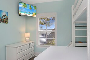 Ein Schlafzimmer mit einem an der Wand montierten Fernseher, einem Fenster mit Jalousien, einem Nachttisch mit Lampe und einem Etagenbett.