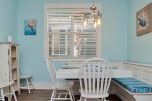Una habitación con un banco blanco, una mesa de comedor y sillas. Hay una ventana con persianas, un cuadro en la pared y una lámpara de araña.