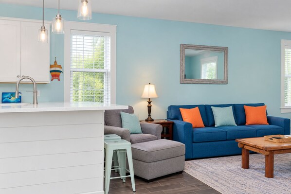 Una sala de estar con un sofá azul, una mesita y una zona de cocina con un lavamanos y luces colgantes.