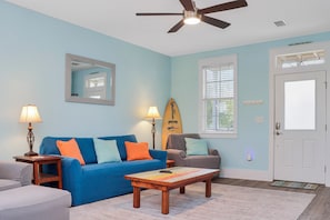 A living room with a blue sofa, two lamps, a surfboard decoration, a ceiling fan, and a door with a window. - Barefoot Cottage A3 (Port St. Joe)
