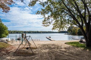 A sandy beach with a swing set, a dock extending into a lake, and boats moored nearby. - Dockside Sands at Sommer Dorf (West Branch)