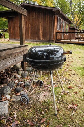 Ein schwarzer Grill mit einem Metallgestell, platziert auf einer Rasenfläche mit Steinen und herabgefallenen Blättern.