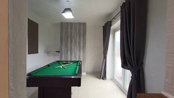 Una mesa de billar en una habitación con ventana, cortinas y una lámpara en el techo.