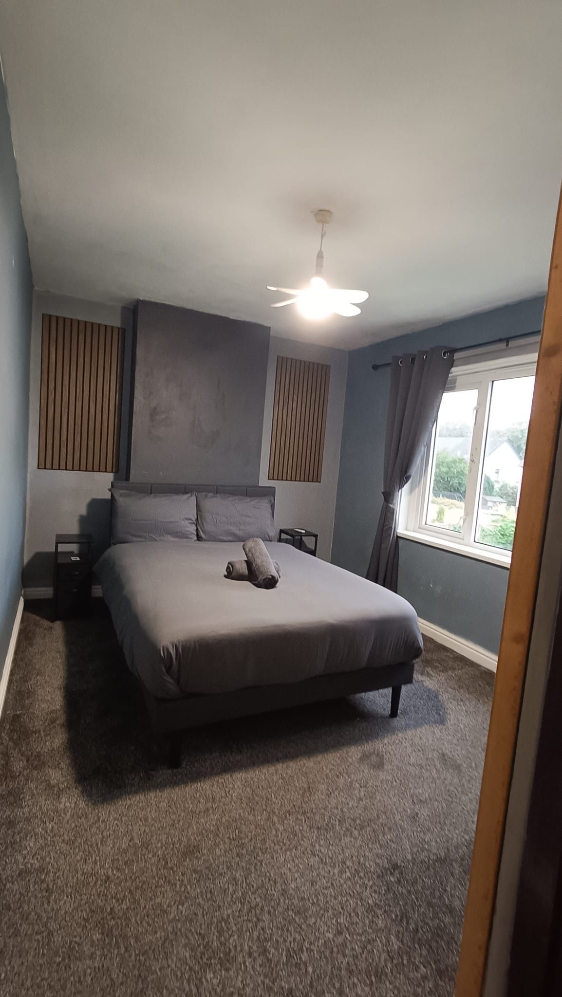 Ein Schlafzimmer mit einem Bett, einem Nachttisch, einem Deckenventilator, einem Fenster mit Vorhängen und Blick ins Freie.