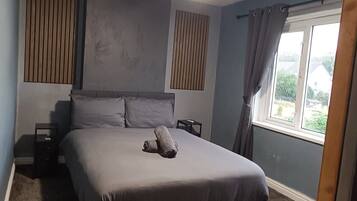 Ein Schlafzimmer mit einem Bett, einem Nachttisch, einem Deckenventilator, einem Fenster mit Vorhängen und Blick ins Freie.