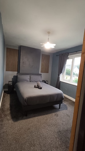 Un dormitorio con cama, mesita de noche, ventilador de techo, ventana con cortinas y vistas al exterior.