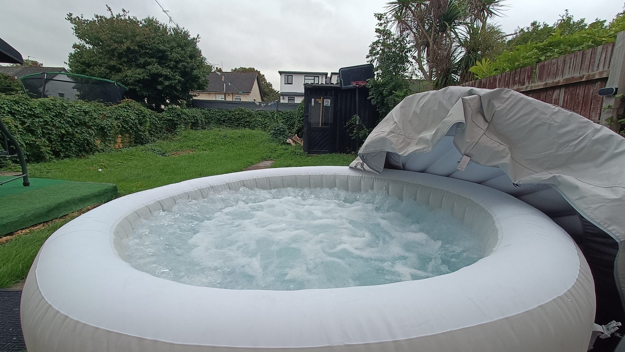 An inflatable hot tub with a cushion and a cover, surrounded by a grassy yard and a wooden fence.