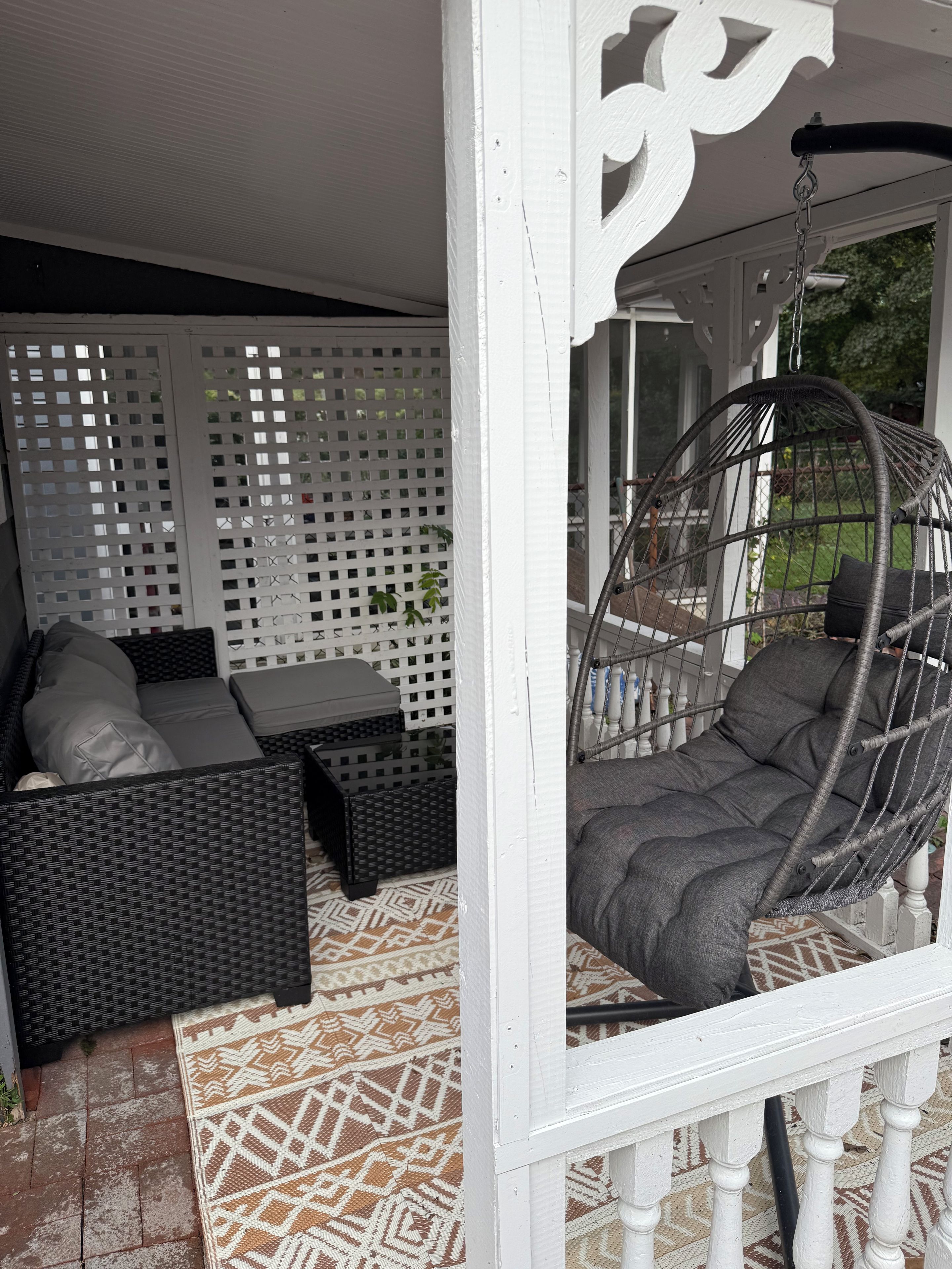 A covered outdoor seating area with a wicker sofa, a hanging chair, and a patterned rug.