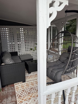 A covered outdoor seating area with a wicker sofa, a hanging chair, and a patterned rug.