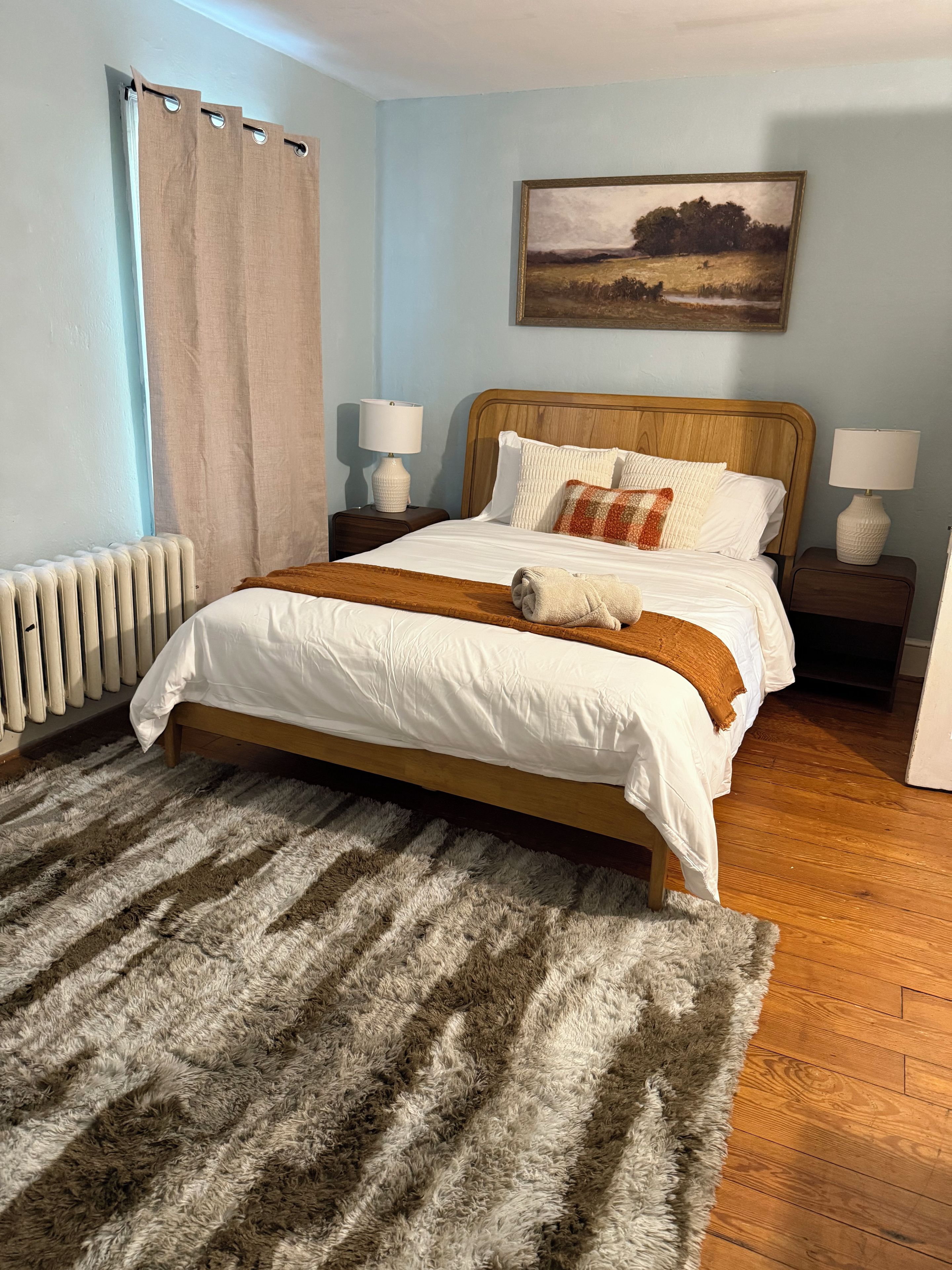 Un dormitorio con cama de madera, ropa de cama blanca, una alfombra mullida, un cuadro en la pared y un radiador.