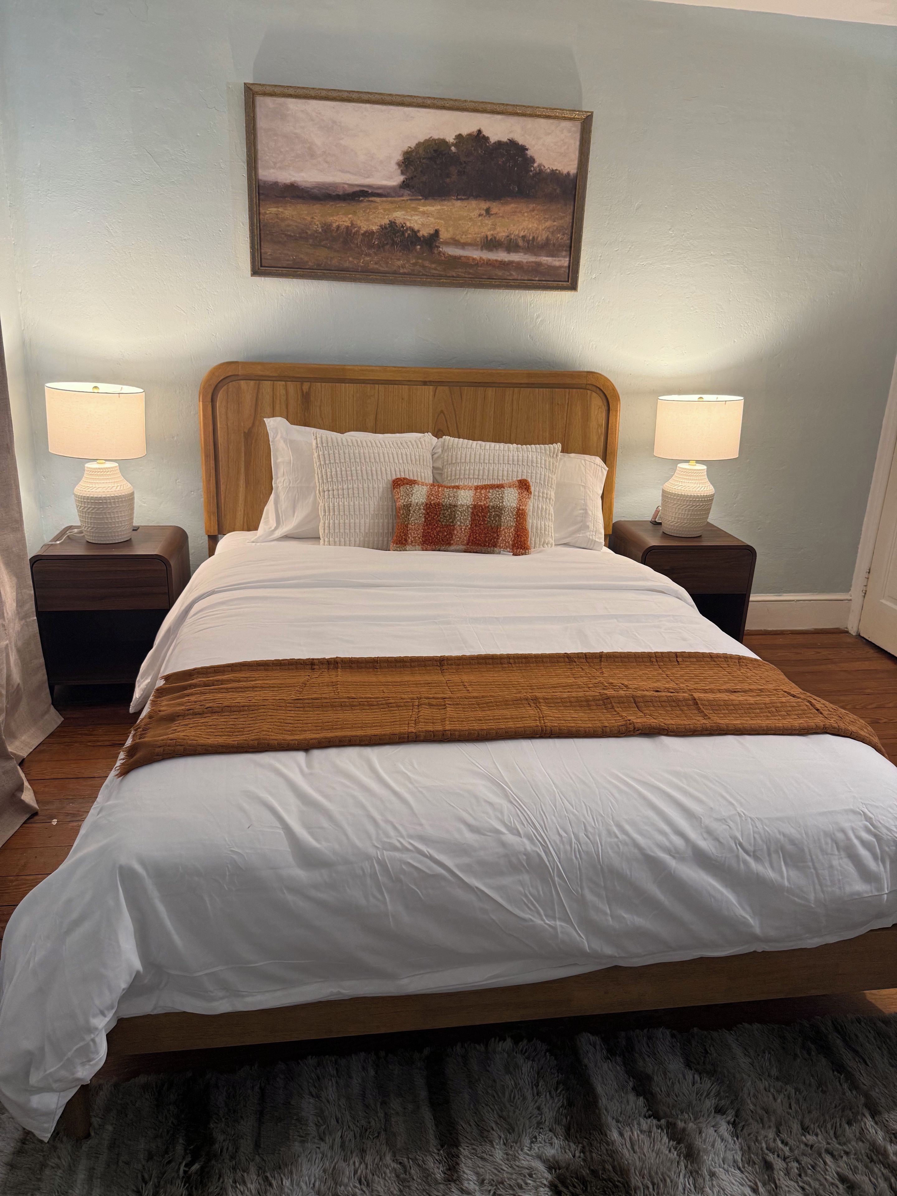 A neatly made bed with white bedding and a brown throw, flanked by two bedside tables with lamps, against a wall with a framed landscape painting.