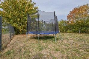 Un trampoline doté d’un filet de sécurité, installé dans un espace extérieur agrémenté d’arbres et entouré d’une clôture.