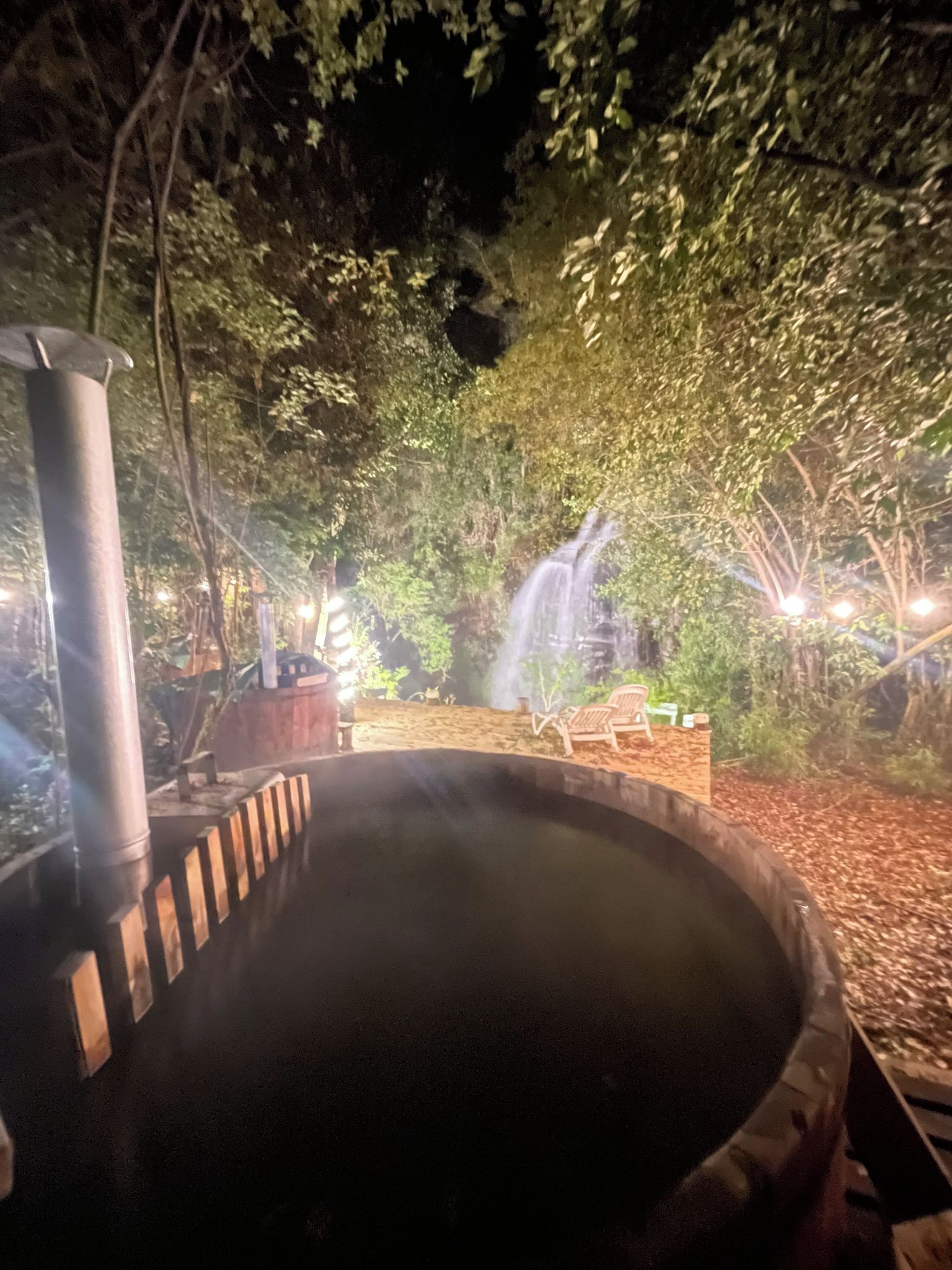 A hot spring area with a wooden deck, a stone pathway, and a waterfall in the background.