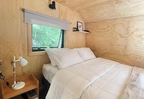 A wooden room with a bed, a nightstand, a desk lamp, a window with a blind, and a shelf with a picture. - Pucura Eco Lodge (Panguipulli)