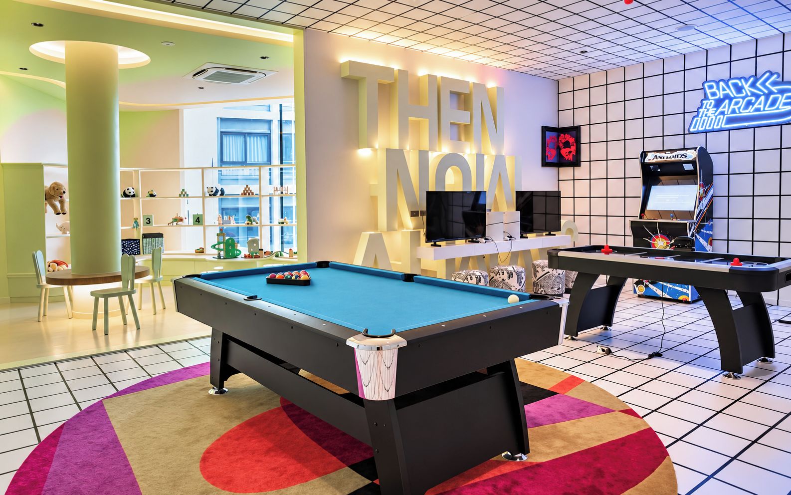 Una mesa de billar con un letrero de neón que dice 'THEN NOW' en una sala con juegos de arcade y una zona de juegos para niños.