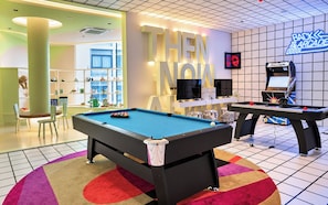 Una mesa de billar con un letrero de neón que dice 'THEN NOW' en una sala con juegos de arcade y una zona de juegos para niños.