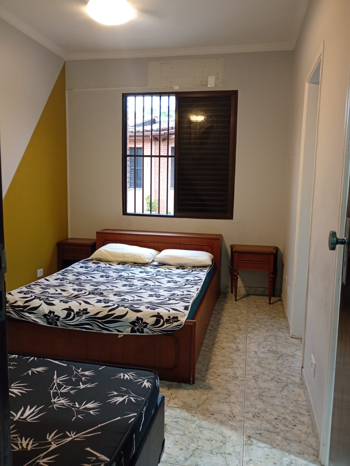Un dormitorio con cama, mesitas de noche, una ventana con barrotes y una puerta.