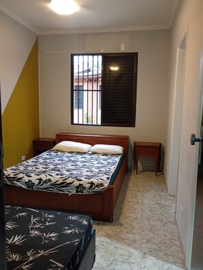 A bedroom with a bed, bedside tables, a window with bars, and a door. - House 50 meters from Enseada beach. (Guarujá)
