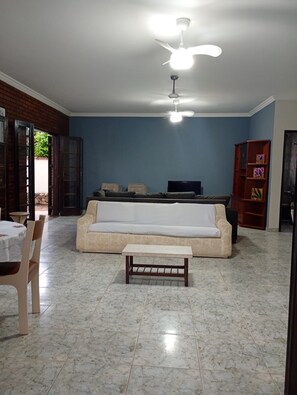 A living room with a sofa, a coffee table, a TV, and a ceiling fan. - House 50 meters from Enseada beach. (Guarujá)