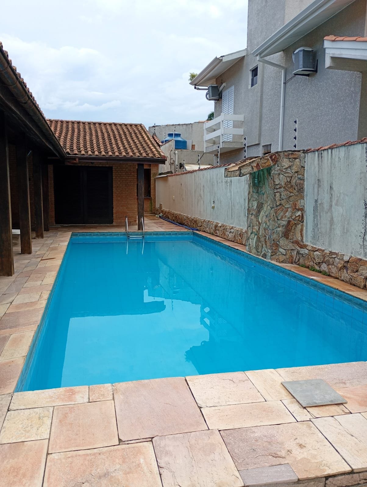 A rectangular swimming pool with clear blue water, surrounded by stone tiles and a stone wall.