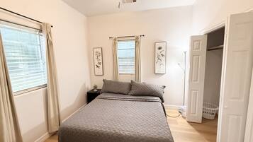 Un dormitorio con cama, ventilador de techo, ventana con cortinas y puerta que da a otra habitación.