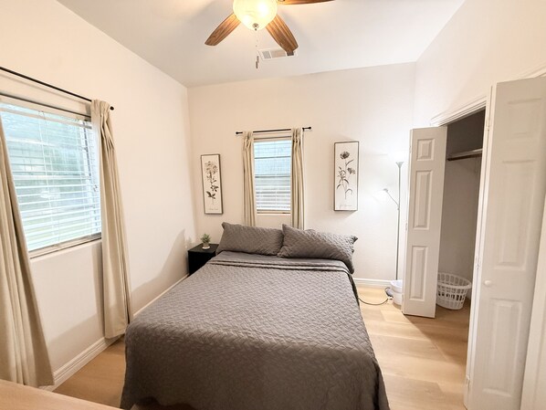 Un dormitorio con cama, ventilador de techo, ventana con cortinas y puerta que da a otra habitación.