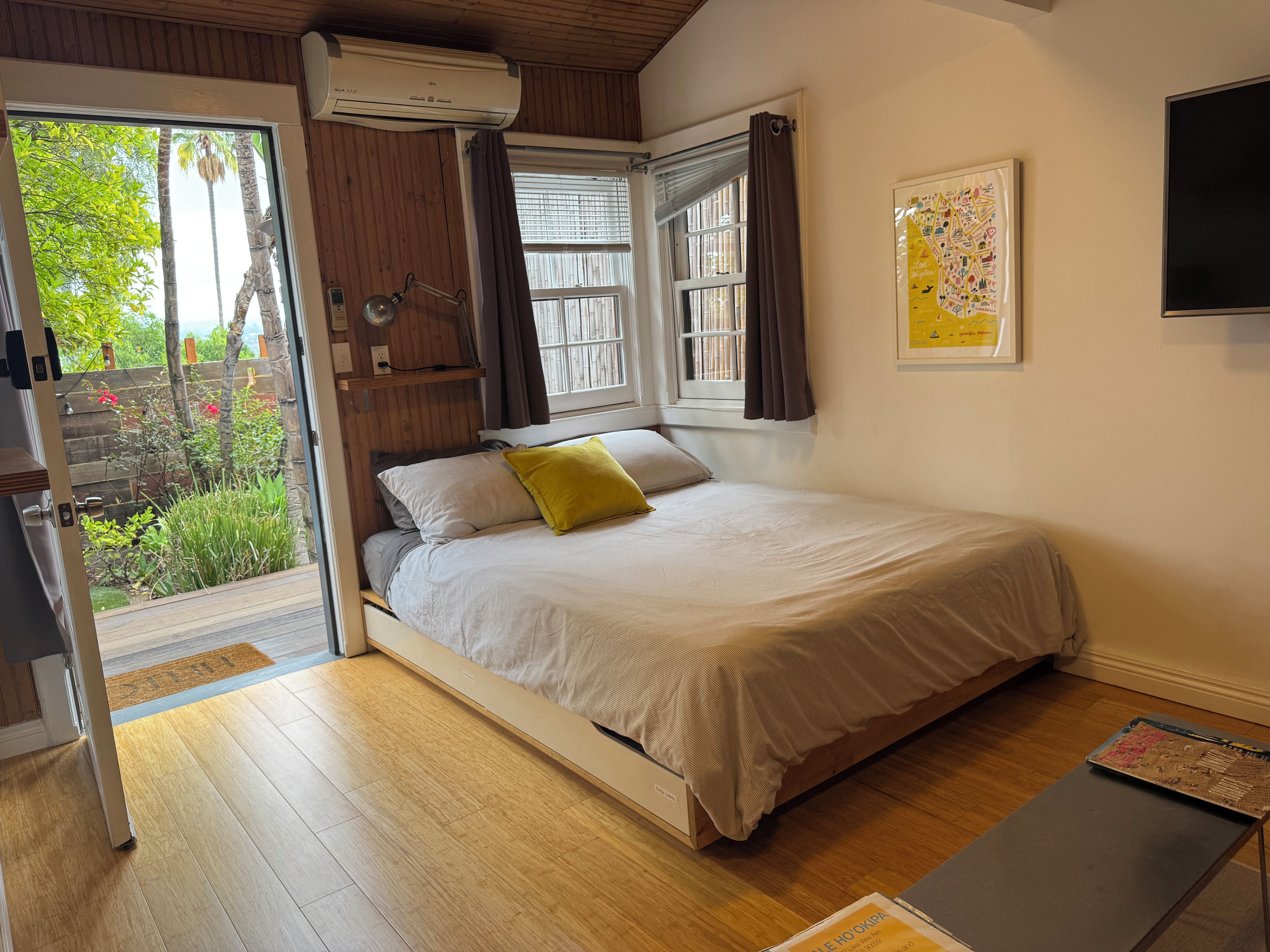 A bedroom with a bed, a window with curtains, a wall-mounted air conditioner, and a door leading to a balcony.