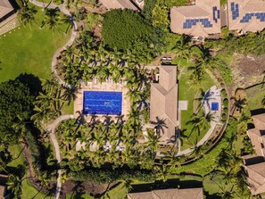 Luchtbeeld van een resort met een centraal zwembad, omringd door palmbomen en begroeiing, en meerdere gebouwen met zonnepanelen.