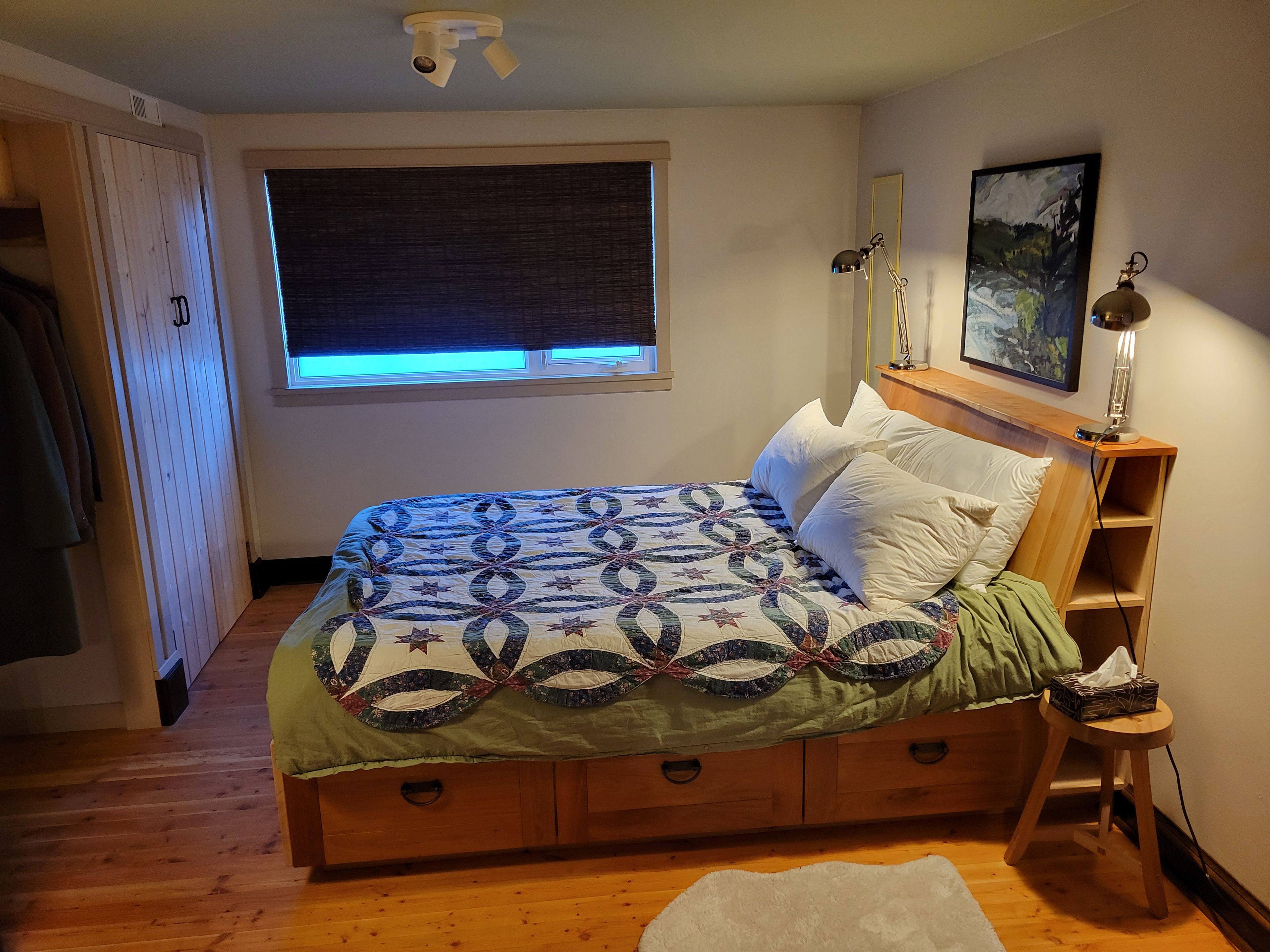 A bedroom with a bed, a window, a nightstand, a lamp, and a painting on the wall.