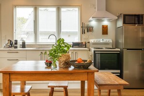 A kitchen with a wooden table, a bowl of fruit, and a plant. - Little Eden by Waiheke Pure (Waiheke Island)