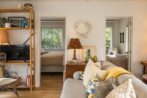 A cozy living room with a sofa, a television, a bookshelf, and a window with a view of greenery.