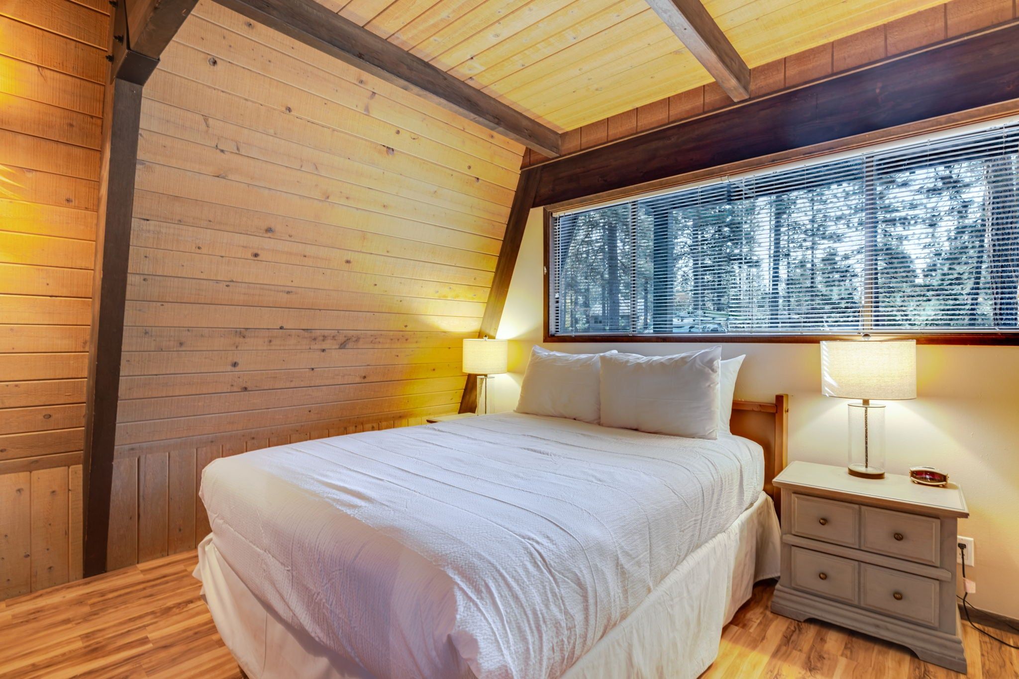 A bedroom with a wooden ceiling, a large window with blinds, a bed with white bedding, two bedside lamps, and a nightstand with a lamp.