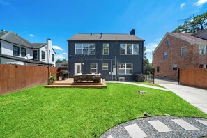 A two-story house with a front yard, a wooden deck, and a brick building in the background. - Spacious 6BR | 2 Homes | Near NRG & Med Center (Houston)