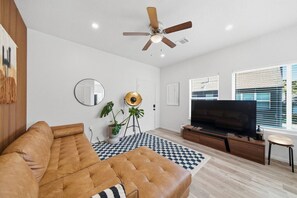 A living room with a brown leather sofa, a TV, a ceiling fan, a large mirror, and a checkered rug. - Spacious 6BR | 2 Homes | Near NRG & Med Center (Houston)