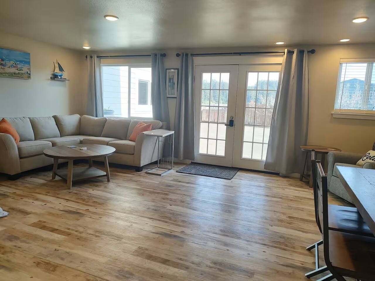 A living room with a sofa, coffee table, and a dining table. There are two windows with curtains, a door, and a view of a deck.