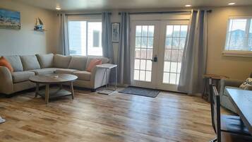 A living room with a sofa, coffee table, and a dining table. There are two windows with curtains, a door, and a view of a deck.