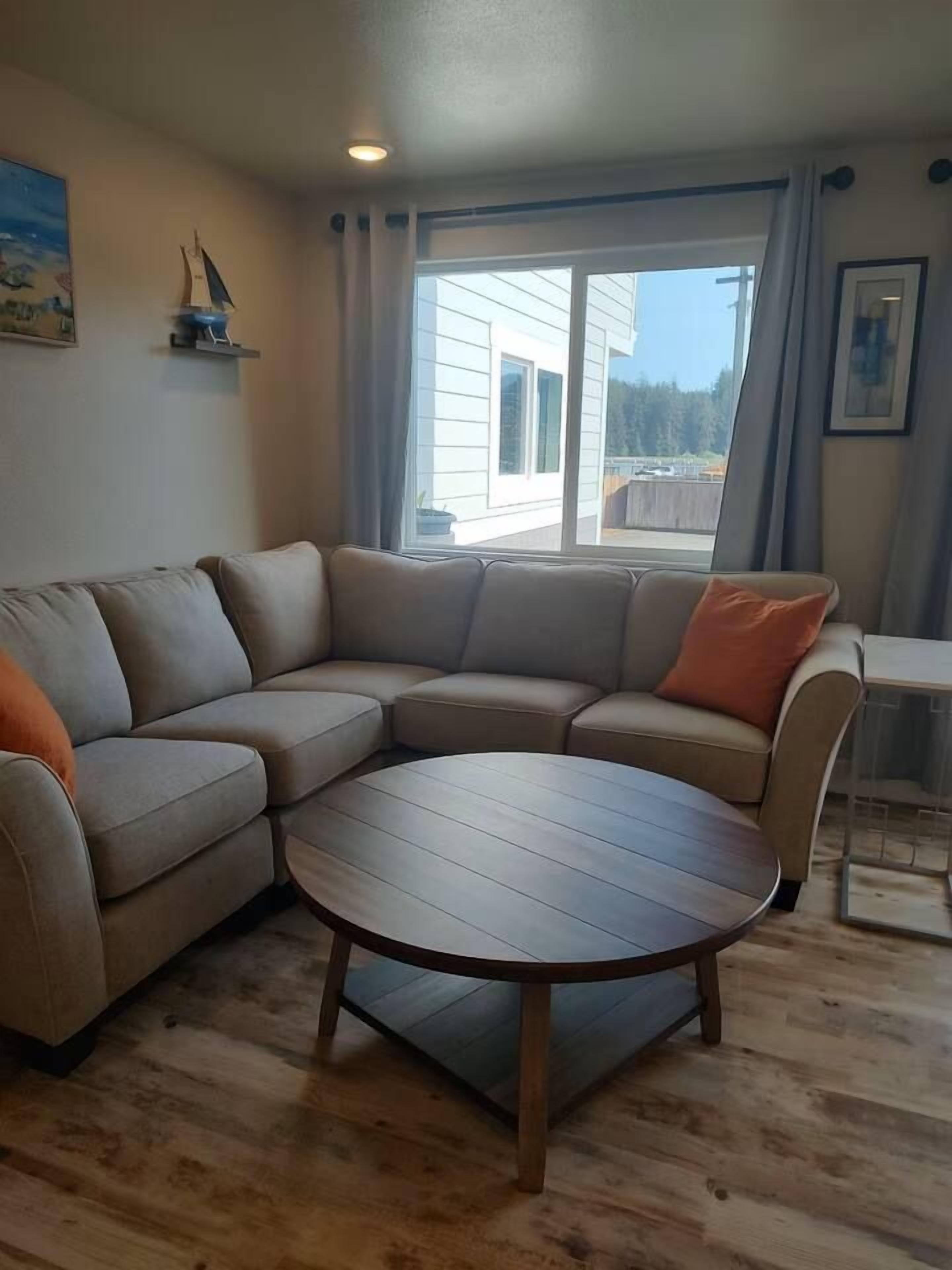 Ett vardagsrum med en hörnsoffa, ett runt soffbord och utsikt över ett hus genom ett fönster.