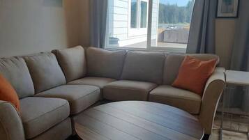 Ett vardagsrum med en hörnsoffa, ett runt soffbord och utsikt över ett hus genom ett fönster.