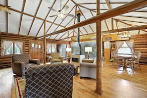 A log cabin interior with a high ceiling, wooden beams, a ceiling fan, a fireplace, and a dining area. - Cedar Crest Cabin: Autumn Views on the Manistee River | Cozy Cabin Getaway (Grayling)