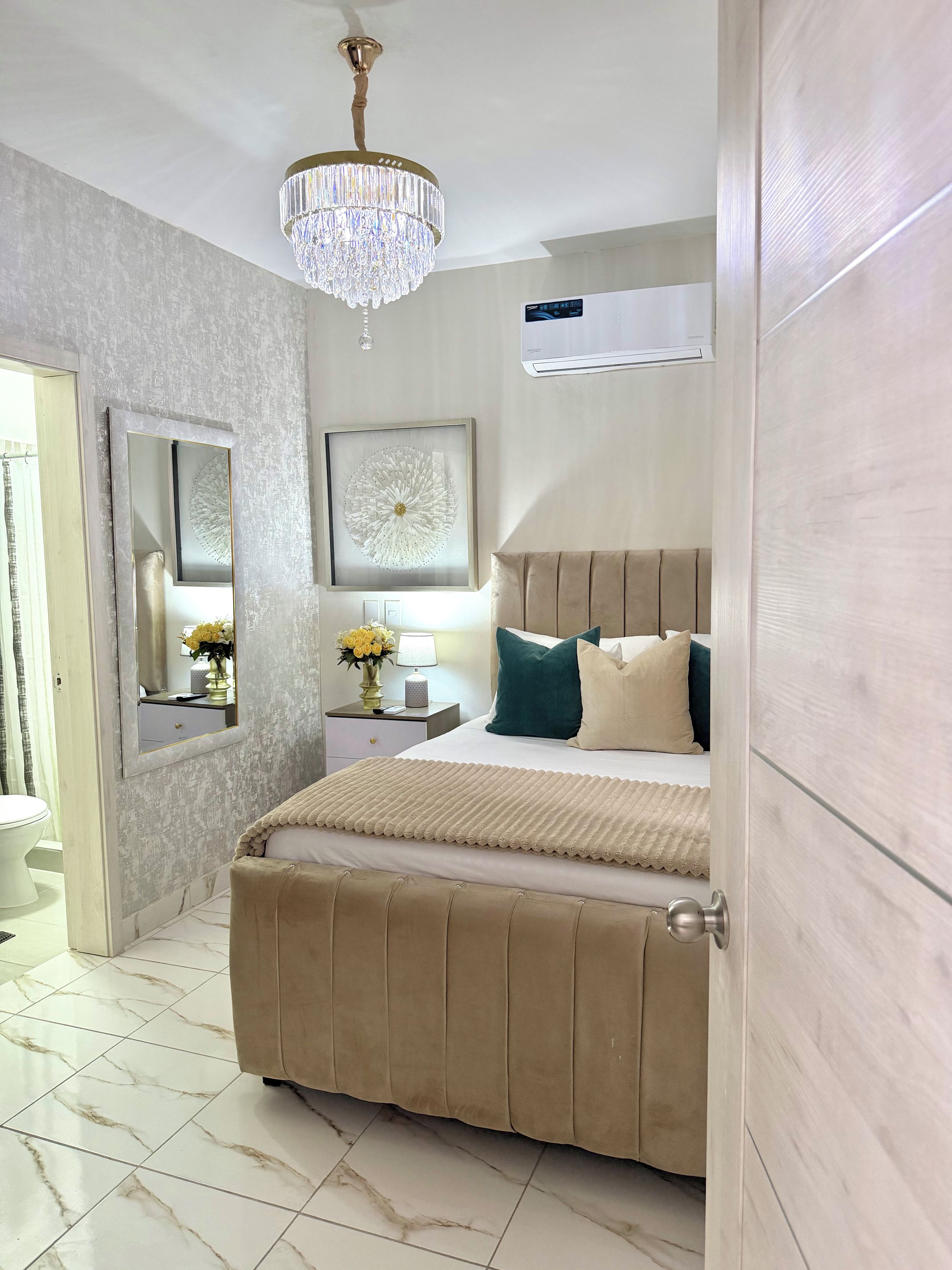 A bedroom with a bed, bedside table, mirror, chandelier, and an air conditioning unit.