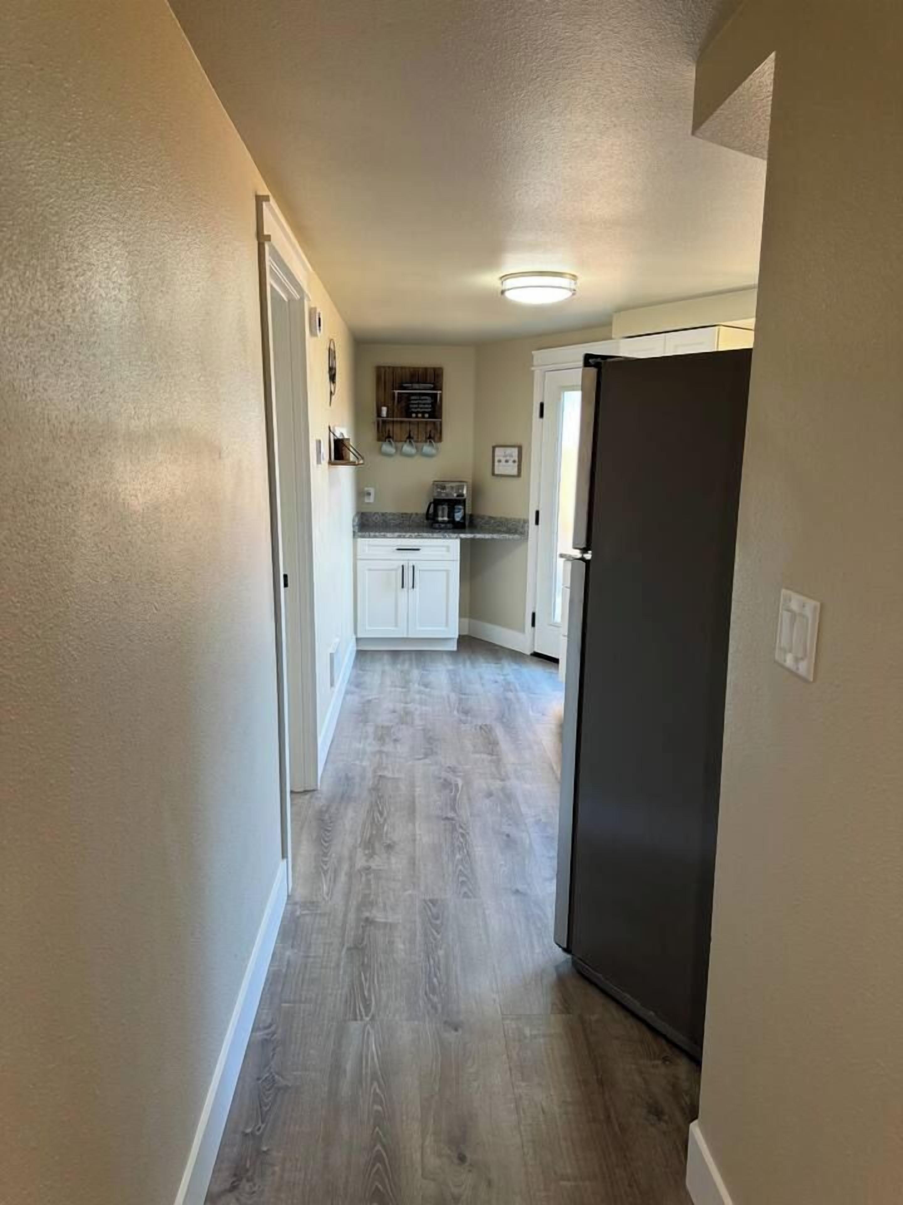 A narrow hallway with a kitchen area at the end, featuring cabinets, a countertop, and a refrigerator.