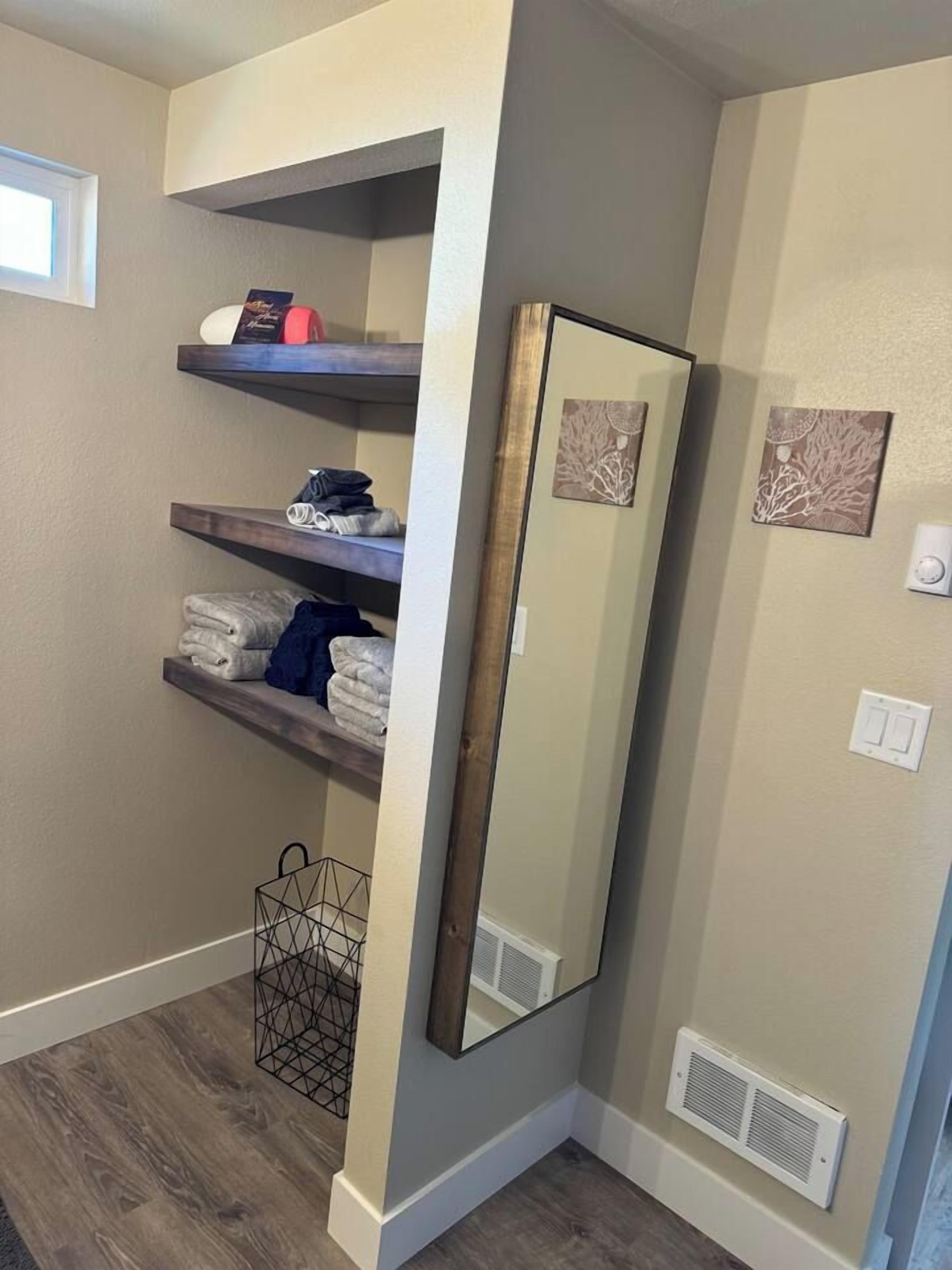 A corner of a room with a built-in shelf unit holding folded towels and a framed mirror.