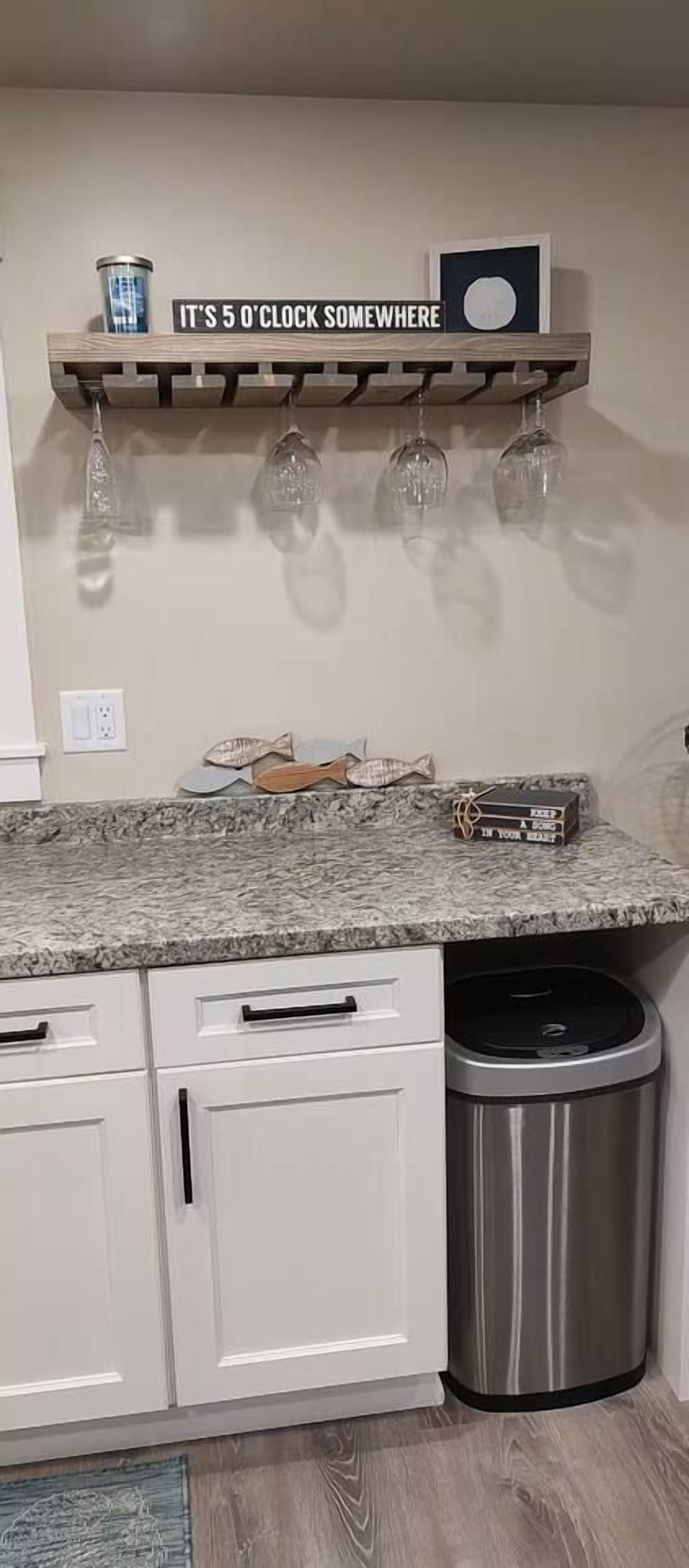 Cozinha com bancada de granito, armários brancos, lixeira de aço inoxidável e prateleira com uma placa e taças de vinho.