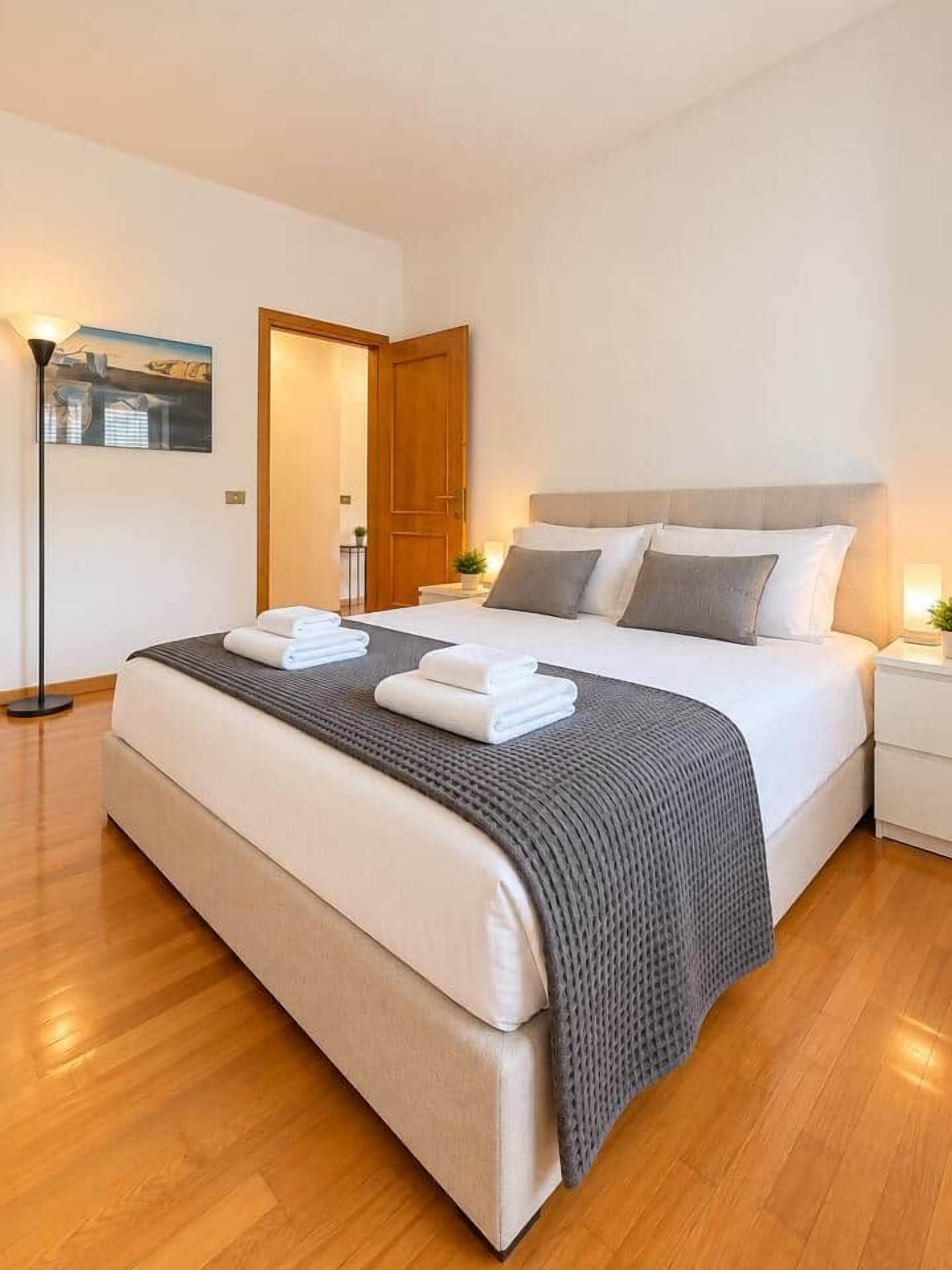 Ein modernes Hotelzimmer mit einem großen Bett, einem Nachttisch, einer Lampe und einem gerahmten Bild an der Wand.