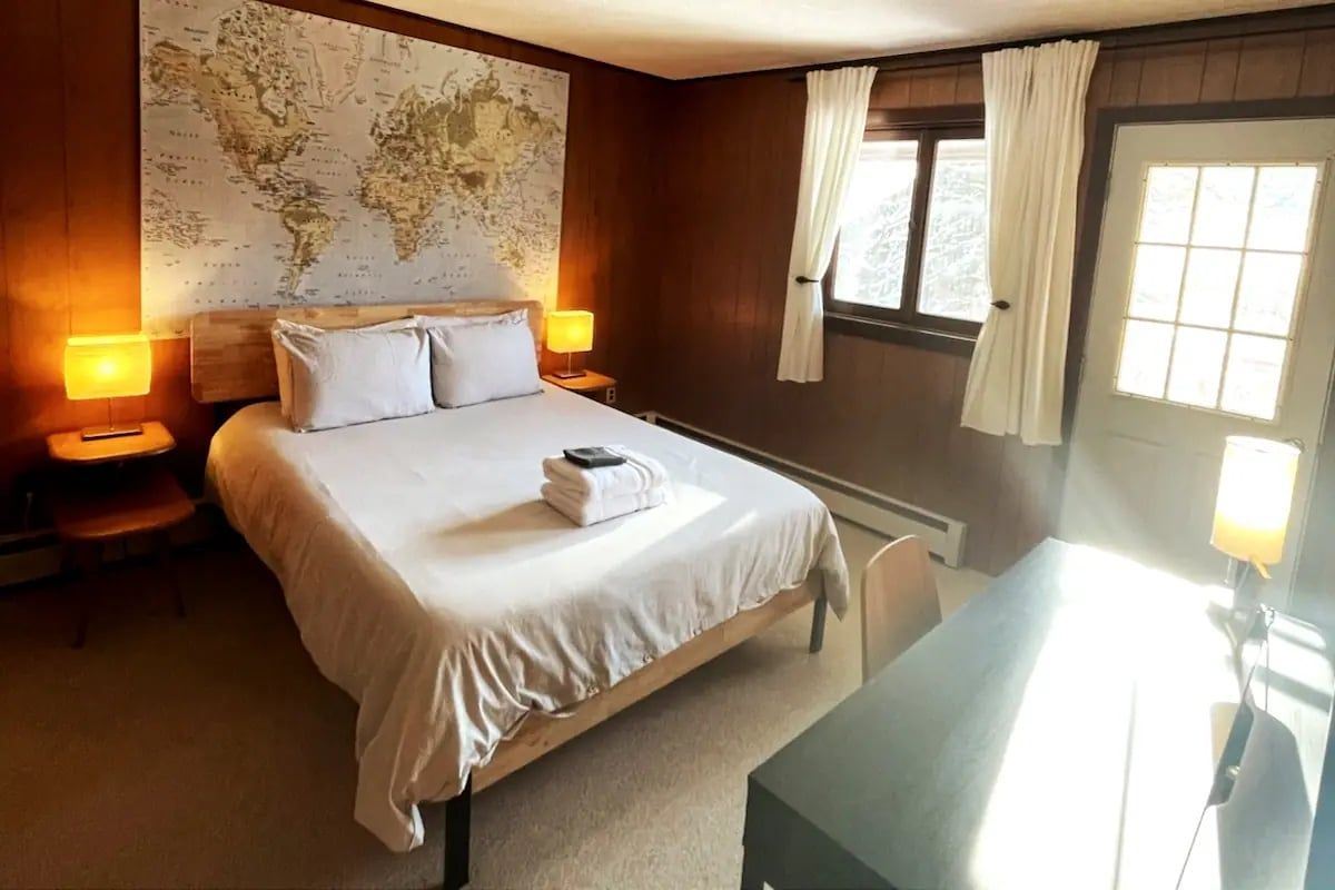 A hotel room with a large bed, two bedside tables with lamps, a world map on the wall, a window with curtains, and a desk.
